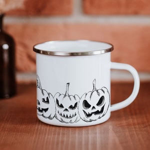 May include: White enamel mug with a black and white illustration of four jack-o'-lantern pumpkins. The mug has a silver rim and handle.