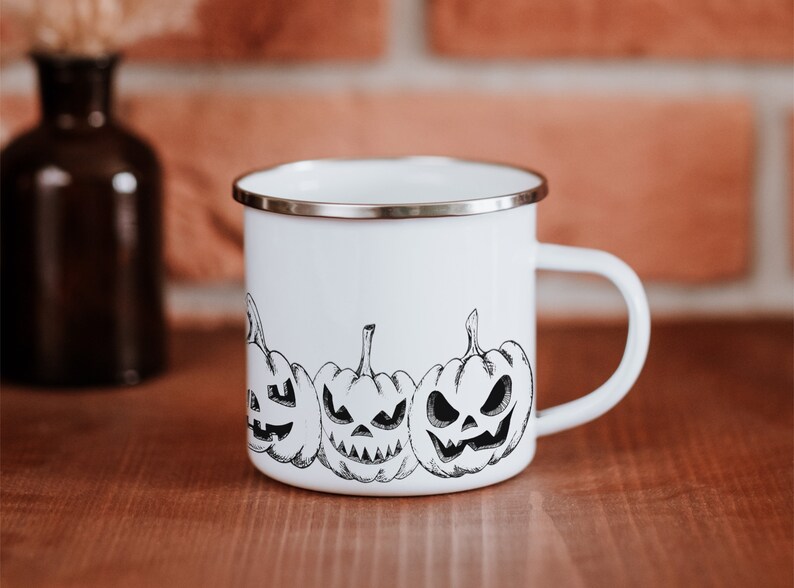 May include: White enamel mug with a black and white illustration of four jack-o'-lantern pumpkins. The mug has a silver rim and handle.