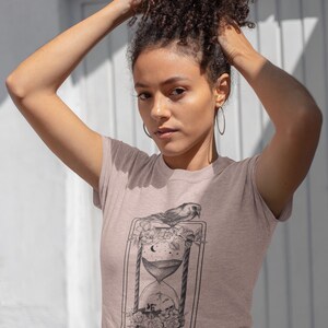May include: A light pink t-shirt with a black and white graphic of an hourglass with a bird perched on top. The graphic is surrounded by flowers and the text "MEMENTO MORI" is printed below the hourglass.