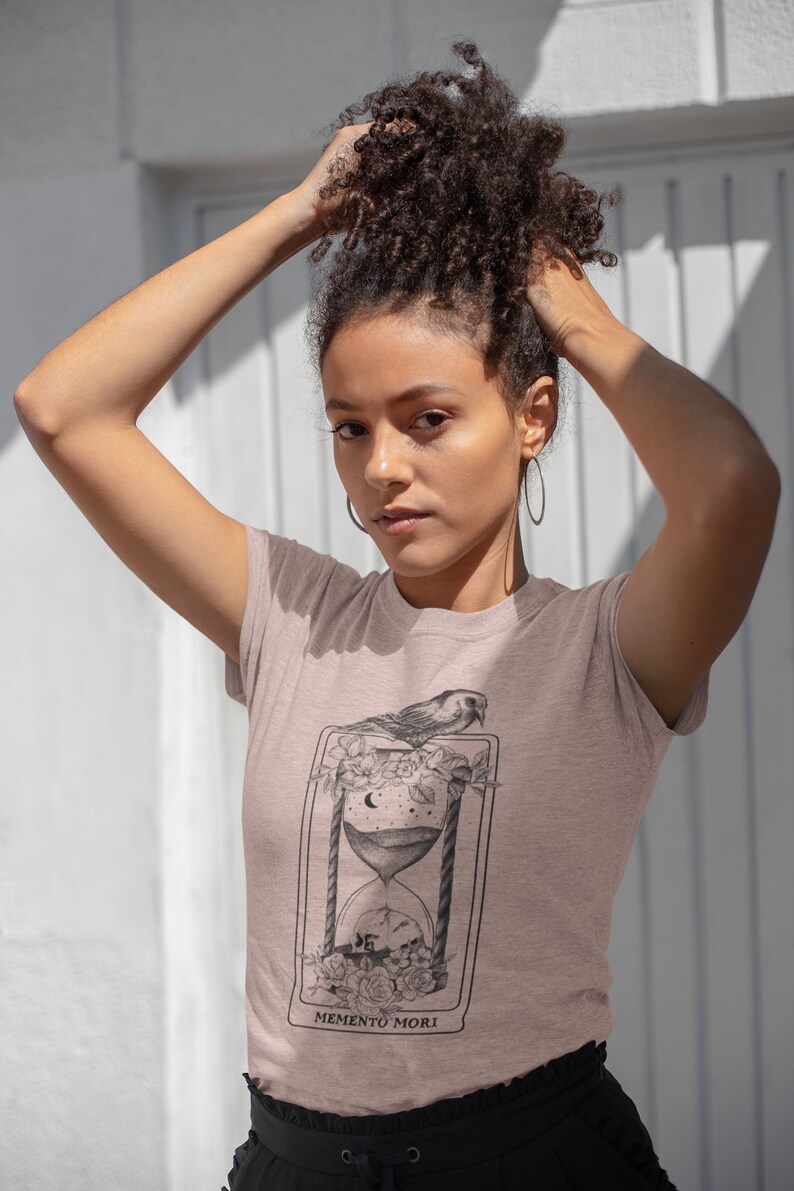 May include: A light pink t-shirt with a black and white graphic of an hourglass with a bird perched on top. The graphic is surrounded by flowers and the text "MEMENTO MORI" is printed below the hourglass.