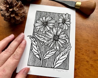 Pode incluir: Uma gravura em linóleo a preto e branco de três margaridas com pétalas e folhas detalhadas. A obra de arte é assinada "M. Soyzeau '25" e numerada "71/100". A impressão é em papel branco, com fundo de madeira e um pinha.