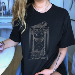 May include: Black t-shirt with a graphic print of an hourglass, a bird, and flowers. The text "Memento Mori" is printed below the graphic.