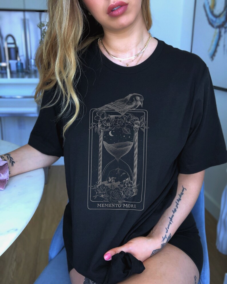 May include: Black t-shirt with a graphic print of an hourglass, a bird, and flowers. The text "Memento Mori" is printed below the graphic.