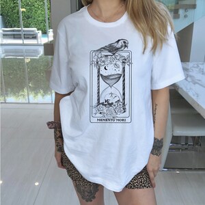 May include: White t-shirt with a black and white graphic of an hourglass, flowers, a bird, and the text "Memento Mori".