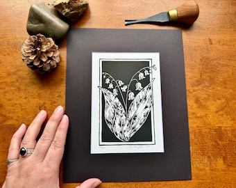 Pode incluir: Impressão em linóleo a preto e branco de flores de lírio-do-vale. A obra de arte é montada em um passe-partout preto. Uma mão com anéis é visível no canto inferior esquerdo. Uma ferramenta de escultura, um pinha e pedras também estão presentes.