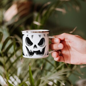 May include: White enamel camping mug with a black and white illustration of a jack-o'-lantern face.