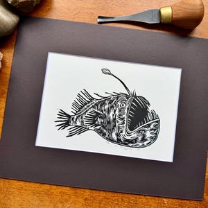 K&ouml;nnte beinhalten: Schwarz-Wei&szlig;-Linolschnitt eines Anglerfischs. Das Kunstwerk befindet sich auf wei&szlig;em Hintergrund, vor einer dunkelgrauen Matte. Ein Holzschnitzwerkzeug und ein Tannenzapfen sind sichtbar. Der Druck zeigt detaillierte Linien.