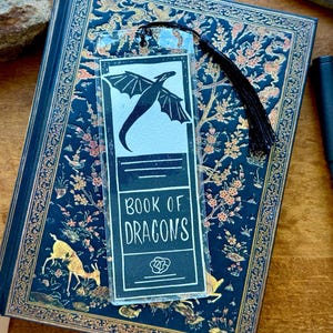 May include: A clear acrylic bookmark featuring a black dragon silhouette and the text "BOOK OF DRAGONS" rests on a dark blue journal with gold floral and animal designs. A black pen lies beside the journal. A black tassel hangs from the bookmark.