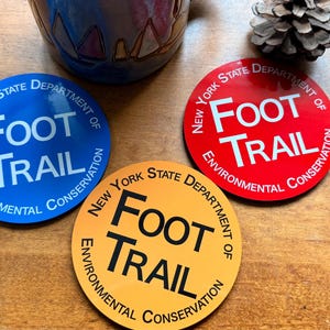 Puede incluir: Tres posavasos circulares en azul, rojo y amarillo. Cada uno presenta el texto "NEW YORK STATE DEPARTMENT OF ENVIRONMENTAL CONSERVATION" arqueado alrededor del borde y las palabras "FOOT TRAIL" impresas en letras grandes y centradas.