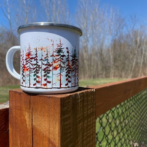 May include: White enamel camping mug with a forest scene design. The mug has a stainless steel lid and a black handle. The mug is sitting on a wooden fence post.