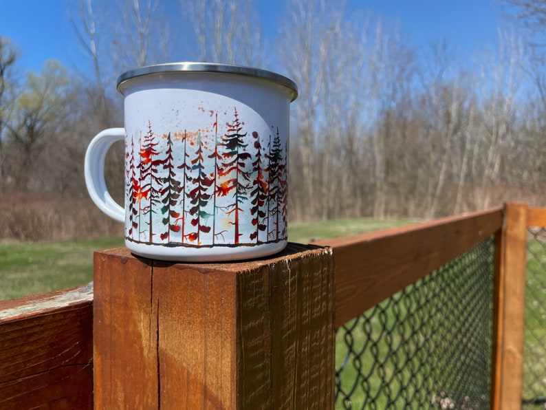 May include: White enamel camping mug with a forest scene design. The mug has a stainless steel lid and a black handle. The mug is sitting on a wooden fence post.