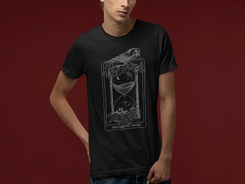 May include: A black t-shirt with a white graphic design of an hourglass with a bird perched on top. The graphic is surrounded by flowers and the text "Memento Mori" is printed below the hourglass.