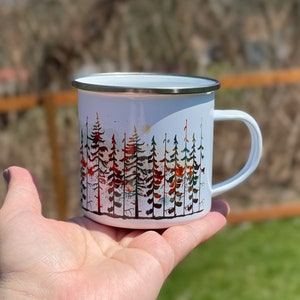 May include: White enamel camping mug with a watercolor design of a forest scene. The trees are in shades of red, orange, green, and blue.