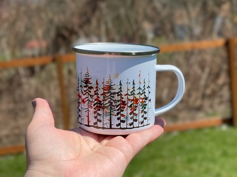 May include: White enamel camping mug with a watercolor design of a forest scene. The trees are in shades of red, orange, green, and blue.