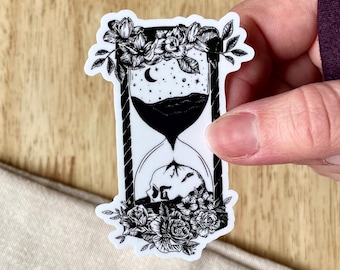 Op de afbeelding: Zwart-witte sticker van een zandloper met een schedel aan de onderkant en bloemen rond de bovenkant. De zandloper is gevuld met zand en sterren.