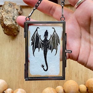 May include: A black silhouette of a dragon with wings outstretched is printed on a white paper background. The paper is framed in a black metal frame with a hanging chain.