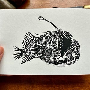 K&ouml;nnte beinhalten: Ein Schwarz-Wei&szlig;-Linolschnitt eines Anglerfisches. Der Fisch hat ein gro&szlig;es Maul, stachelige Flossen und einen leuchtenden K&ouml;der. Der Druck ist auf strukturiertem, wei&szlig;em Papier. Vom K&uuml;nstler signiert.