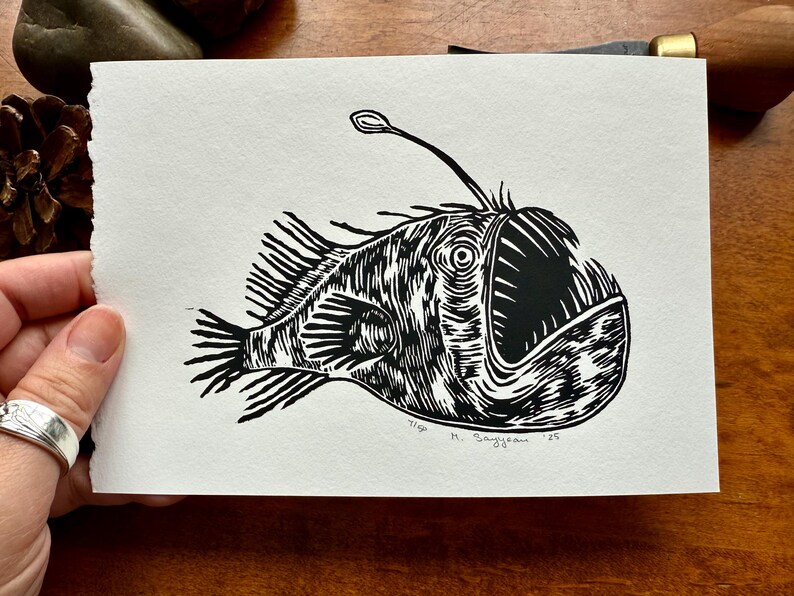 K&ouml;nnte beinhalten: Ein Schwarz-Wei&szlig;-Linolschnitt eines Anglerfisches. Der Fisch hat ein gro&szlig;es Maul, stachelige Flossen und einen leuchtenden K&ouml;der. Der Druck ist auf strukturiertem, wei&szlig;em Papier. Vom K&uuml;nstler signiert.