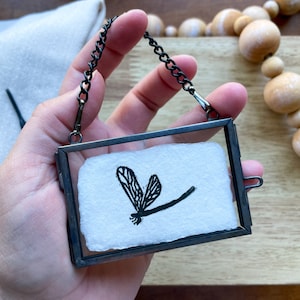 Può includere: Una piccola cornice rettangolare in metallo nero con una catena, che contiene un pezzo di carta bianca con un'illustrazione di libellula nera. La libellula è raffigurata con ali dettagliate e un corpo sottile. La cornice è tenuta da una mano.