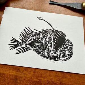 K&ouml;nnte beinhalten: Schwarz-Wei&szlig;-Linolschnitt eines Anglerfisches. Der Druck zeigt detaillierte Linien, die die einzigartigen Merkmale des Fisches hervorheben, wie sein gro&szlig;es Maul und den K&ouml;der. Vom K&uuml;nstler signiert, der Druck befindet sich auf strukturiertem Papier.