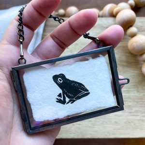 Pode incluir: Pingente retangular com moldura de metal preto. Dentro da moldura há um pedaço de papel branco com uma ilustração de sapo preto. O pingente pende de uma corrente preta. O fundo inclui contas de madeira.