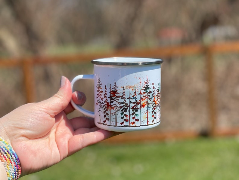 May include: White enamel mug with a colorful watercolor design of a forest. The trees are in shades of red, orange, yellow, green, and blue.