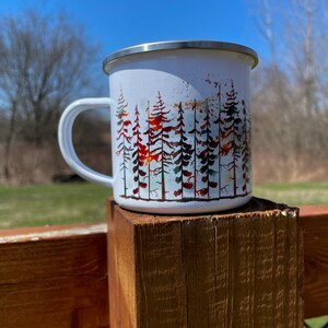 May include: White enamel camping mug with a watercolor design of a forest of trees in shades of green, red, and brown.