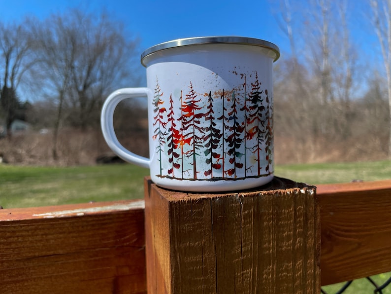 May include: White enamel camping mug with a watercolor design of a forest of trees in shades of green, red, and brown.