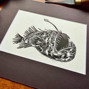 K&ouml;nnte beinhalten: Schwarz-Wei&szlig;-Linolschnitt eines Anglerfischs. Das Kunstwerk zeigt eine detaillierte Illustration des Tiefsee-Kreaturs mit seinem charakteristischen K&ouml;der. Der Druck ist mattiert und gerahmt, fertig zum Aufh&auml;ngen. Die Signatur des K&uuml;nstlers und das Jahr sind sichtbar.
