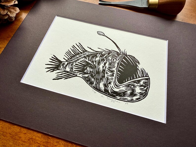 K&ouml;nnte beinhalten: Schwarz-Wei&szlig;-Linolschnitt eines Anglerfischs. Das Kunstwerk zeigt eine detaillierte Illustration des Tiefsee-Kreaturs mit seinem charakteristischen K&ouml;der. Der Druck ist mattiert und gerahmt, fertig zum Aufh&auml;ngen. Die Signatur des K&uuml;nstlers und das Jahr sind sichtbar.
