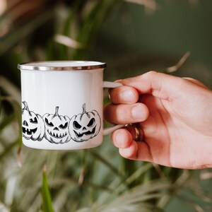 May include: White enamel mug with a black and white illustration of three jack-o'-lantern faces.