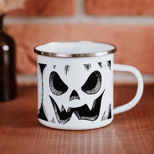 May include: White enamel mug with a black and white illustration of a jack-o'-lantern face.