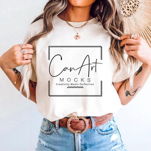 以下が含まれることがあります： 「CanArt Mocks」というテキストと「Creativity Meets Perfection」というテキストが黒い正方形で囲まれた白いTシャツ。モデルは、茶色のベルトと薄いブルーのデニムのショートパンツを着用してTシャツを着ています。
