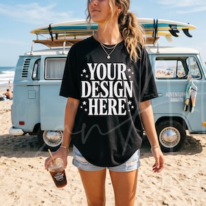 May include: Black t-shirt with the text "YOUR DESIGN HERE" in white, with a beach scene in the background. A light blue van with surfboards on top is parked on the sand. The person is holding a coffee drink.