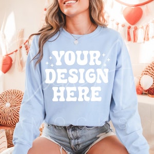 Puede incluir: Una sudadera de color azul claro con el texto "YOUR DESIGN HERE" en blanco. La sudadera está siendo modelada por una persona que lleva pantalones cortos de mezclilla.