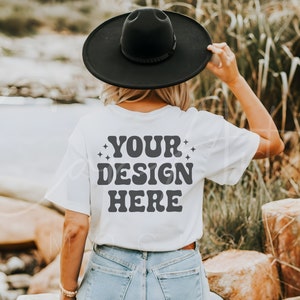 以下が含まれることがあります： レトロなフォントで「YOUR DESIGN HERE」と書かれた白いTシャツ。黒い帽子と青いジーンズを着用した人が着ています。