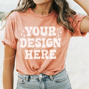 May include: A woman wearing a peach-colored t-shirt with the text "YOUR DESIGN HERE" printed in white. The shirt is tied at the waist and the woman is wearing blue jeans.