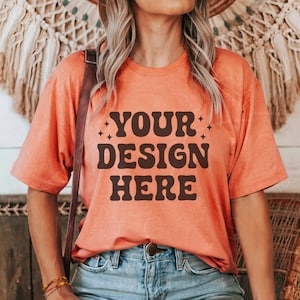 Puede incluir: Una camiseta de color naranja coral con mangas cortas y cuello redondo. La camiseta tiene el texto "YOUR DESIGN HERE" impreso en marrón en la parte delantera.