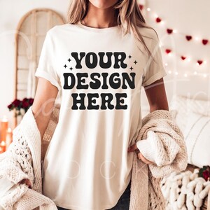 May include: A white t-shirt with the text "YOUR DESIGN HERE" in black lettering. The shirt is being worn by a person in a cozy setting with string lights and a blanket.