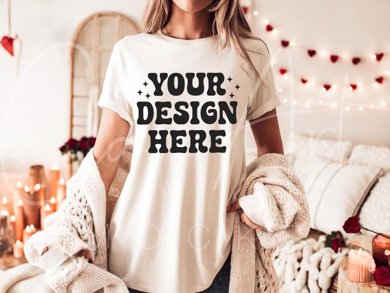 May include: A white t-shirt with the text "YOUR DESIGN HERE" in black lettering. The shirt is being worn by a person in a cozy setting with string lights and a blanket.