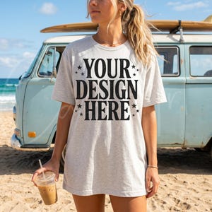 May include: A light gray t-shirt with the text "YOUR DESIGN HERE" in black, with star accents. The shirt is worn by a person on a beach, with a vintage blue van and surfboard in the background. The person is holding a clear plastic cup with a brown beverage.