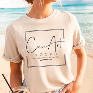 May include: A woman wearing a beige t-shirt with the text "Can Art Mocks Creativity Meets Perfection" printed on it. She is standing on a beach and holding a glass of iced coffee.