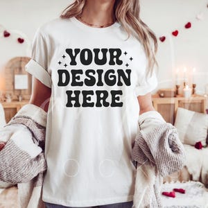 May include: A white t-shirt with the text "YOUR DESIGN HERE" in black lettering. The shirt is being worn by a person in a casual setting.