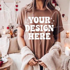 May include: A brown t-shirt with the text "YOUR DESIGN HERE" in white. The shirt is being worn by a person in a cozy setting with candles and blankets.