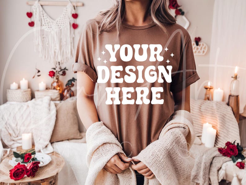 May include: A brown t-shirt with the text "YOUR DESIGN HERE" in white. The shirt is being worn by a person in a cozy setting with candles and blankets.