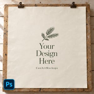 Peut inclure: Une feuille de papier blanc cassé carrée fixée par deux pinces métalliques sur une planche en bois. Le papier présente un dessin au trait vert foncé d'une branche de pin, le texte Your Design Here et la mention CanArtMockups.