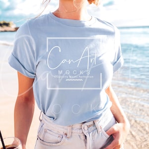 May include: A woman wearing a light blue t-shirt with the text "CanArt" and "MOCKS" in white. The text below "MOCKS" reads "Creativity Meets Perfection". She is wearing light blue denim shorts and holding a glass of iced coffee.