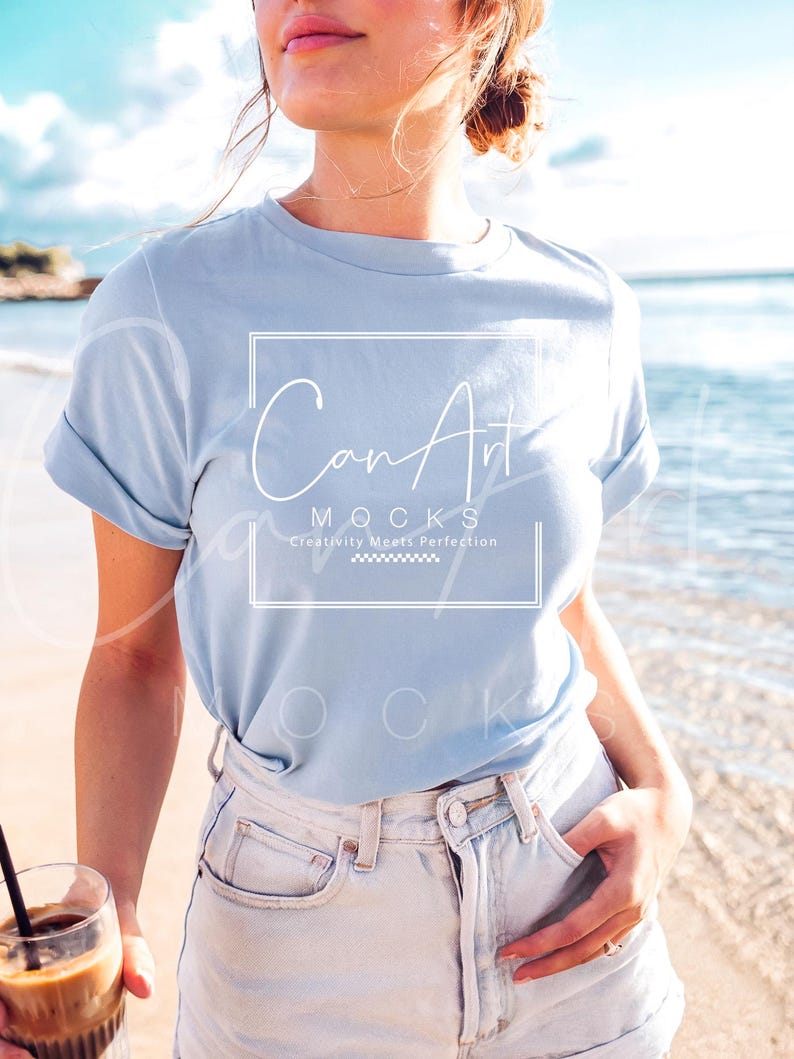 May include: A woman wearing a light blue t-shirt with the text "CanArt" and "MOCKS" in white. The text below "MOCKS" reads "Creativity Meets Perfection". She is wearing light blue denim shorts and holding a glass of iced coffee.