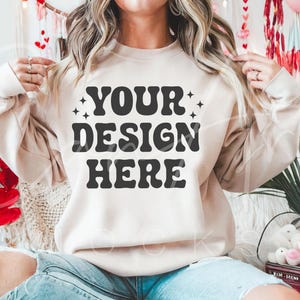 Puede incluir: Una sudadera de color crema con el texto "YOUR DESIGN HERE" en negro. La sudadera está siendo modelada por una persona que lleva pantalones vaqueros azules.