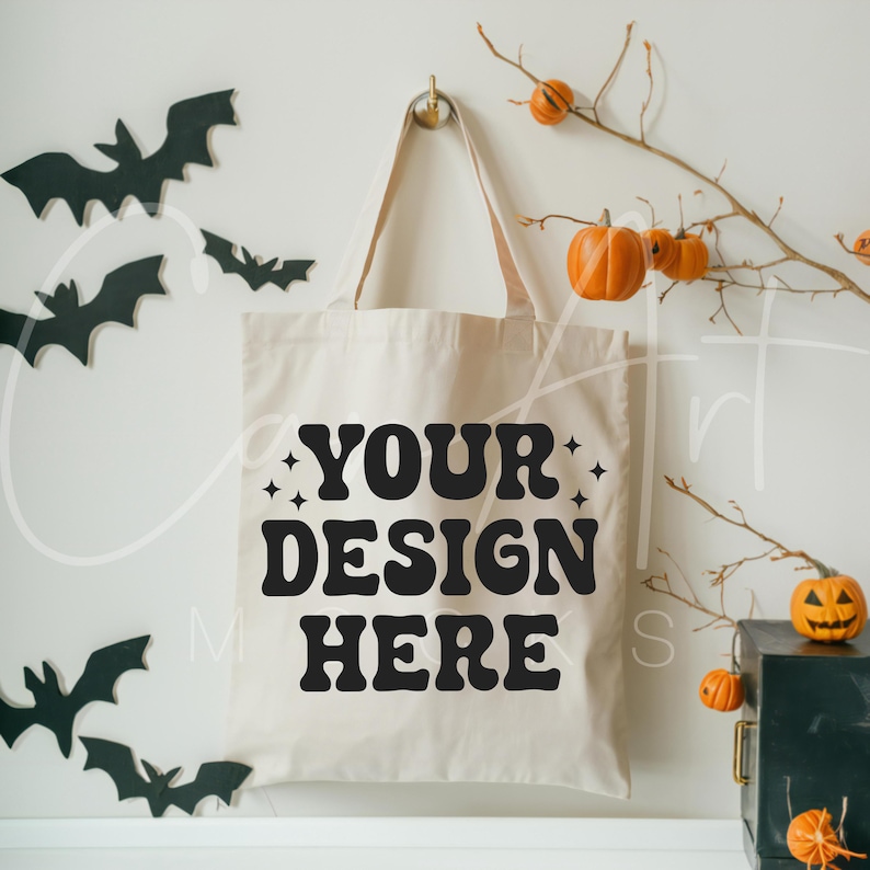 May include: A white canvas tote bag with black text that says "YOUR DESIGN HERE" and stars on either side of the text. The bag is hanging on a white wall with a branch of fall leaves and pumpkins behind it.